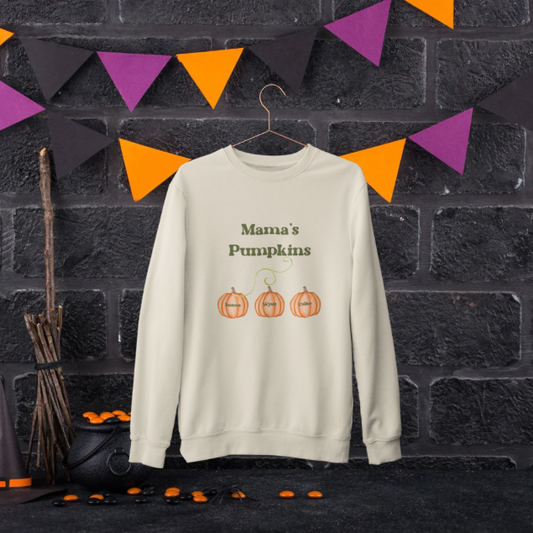 Cream crewneck custom Mama’s Pumpkins sweatshirt hanging on black brick wall with Halloween bunting and three pumpkin graphics