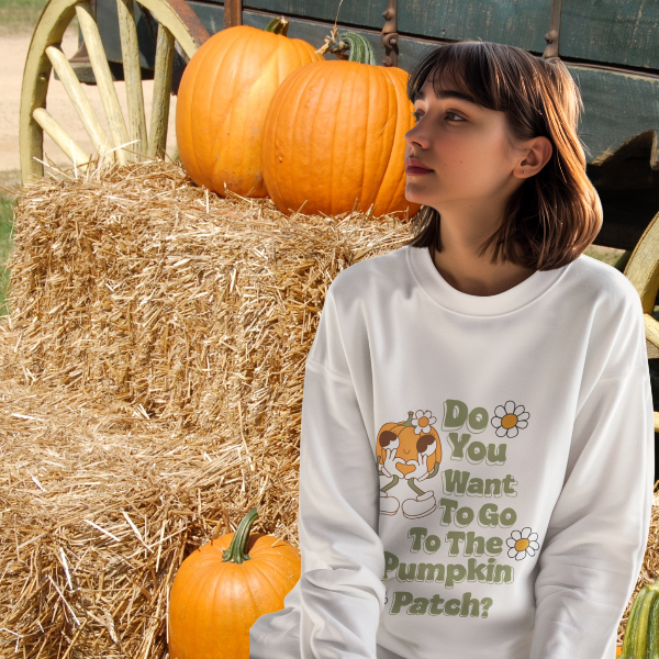 Woman wearing a white Pumpkin Patch sweatshirt with retro daisy design sitting on hay bales by pumpkins